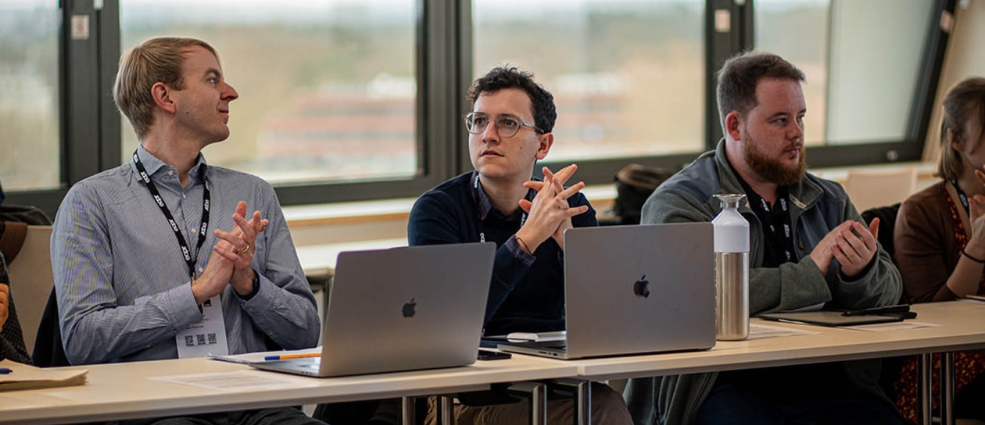 Three people sit at a conference table with laptops, listening and clapping during a meeting. Large windows are in the background.